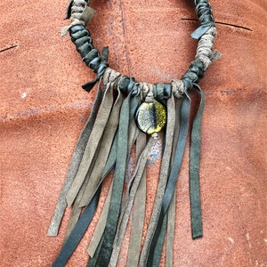 Puede incluir: Un collar de cuero verde y marrón con un colgante redondo de vidrio amarillo y verde. El collar tiene una franja de hebras de cuero.