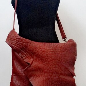 Pu&ograve; includere: Una borsa a tracolla in pelle rossa con una superficie strutturata. La borsa ha una lunga tracolla e una chiusura a lembo.
