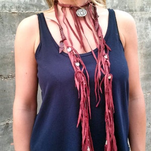 Puede incluir: Un collar largo de cuero burdeos con flecos y detalles plateados. El collar está en capas y tiene un gran colgante circular.