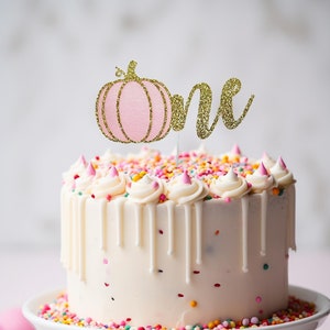 May include: A white frosted cake with colorful sprinkles and a gold glitter "one" cake topper featuring a pink pumpkin. The cake sits on a white cake stand, with a pink background.