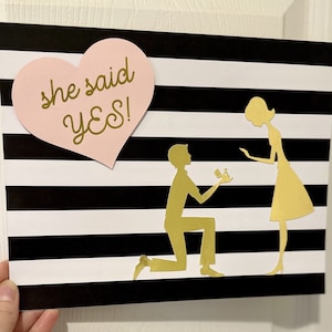 K&ouml;nnte beinhalten: Ein schwarz-wei&szlig; gestreifter Hintergrund mit einem rosa Herz, auf dem "she said yes!" steht. Zwei goldene Silhouetten von Personen befinden sich auf dem Hintergrund. Die Frau steht und der Mann kniet mit einem Ring in der Hand.