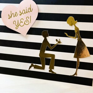 K&ouml;nnte beinhalten: Eine schwarz-wei&szlig; gestreifte Karte mit einem rosa Herz, auf dem "she said yes!" steht. Auf der Karte ist eine goldene Silhouette eines Paares zu sehen. Der Mann kniet und h&auml;lt eine Ringbox in der Hand. Die Frau steht und schaut auf die Ringbox.