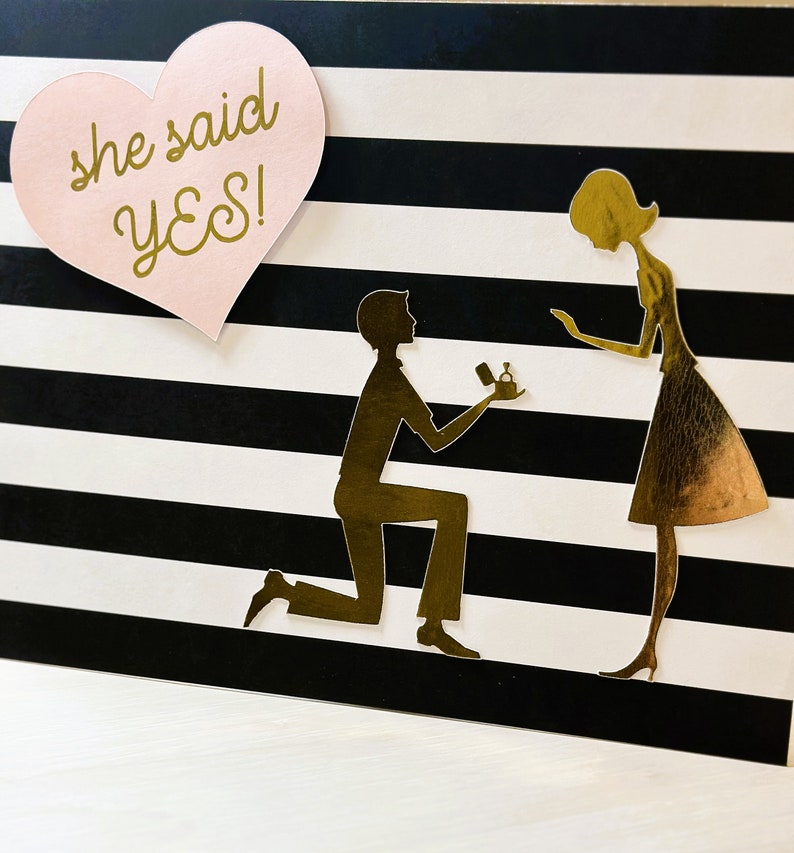 K&ouml;nnte beinhalten: Eine schwarz-wei&szlig; gestreifte Karte mit einem rosa Herz, auf dem "she said yes!" steht. Auf der Karte ist eine goldene Silhouette eines Paares zu sehen. Der Mann kniet und h&auml;lt eine Ringbox in der Hand. Die Frau steht und schaut auf die Ringbox.