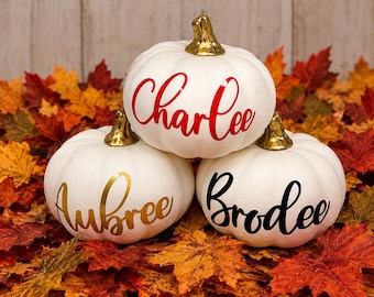 Puede incluir: Tres calabazas decorativas blancas con tallos dorados metálicos. La calabaza superior presenta el nombre Charlee en letra roja. La calabaza inferior izquierda muestra el nombre Aubree en dorado, y la inferior derecha el nombre Brodee en color negro.