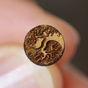May include: A small, round wooden disc with a laser-etched design of a tree with roots and branches. The tree is in the shape of a yin-yang symbol.