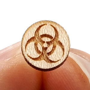 May include: A small, round wooden button with a laser-etched biohazard symbol. The button is light brown in color and features a three-lobed design within a circle. The button is held between two fingers against a white background.