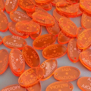 Peut inclure: Un grand tas de breloques ovales en acrylique orange avec le texte "ZAZA BaZOOKA" imprimé dessus. Les breloques ont un trou en haut pour les attacher à un collier ou un bracelet.