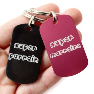 Puede incluir: Dos llaveros negros y rojos con anillos plateados. El llavero negro tiene el texto "super parrain" y el llavero rojo tiene el texto "super marraine".
