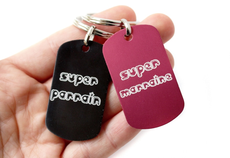 Puede incluir: Dos llaveros negros y rojos con anillos plateados. El llavero negro tiene el texto "super parrain" y el llavero rojo tiene el texto "super marraine".