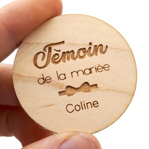 Peut inclure: Une étiquette ronde en bois avec le texte "Temoin de la mariée" et un symbole de nœud papillon, suivi du nom "Coline".
