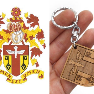 Peut inclure: Un porte-clés en bois représentant un écusson avec un oiseau et des clés, inspiré d'un blason. L'écu est brun avec un oiseau brun clair et des clés dorées. Le texte "Nomen Est Omen" est inscrit sur l'écu.