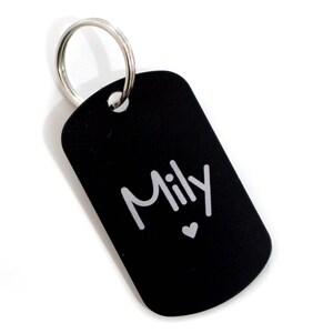 Puede incluir: Chapa de perro de metal negro con un anillo plateado y el nombre "Mily" grabado en ella con un peque&ntilde;o coraz&oacute;n.