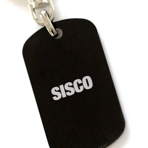 Puede incluir: Llavero de metal negro con un anillo plateado y una cadena plateada. La etiqueta tiene la palabra "SISCO" impresa en blanco.