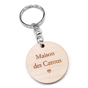 Peut inclure: Un porte-clés en bois avec un anneau et une chaîne en métal argenté. Le pendentif rond en bois porte l'inscription "Maison des Catous" gravée, ainsi qu'un petit symbole de cœur. Le porte-clés est posé sur un fond blanc.