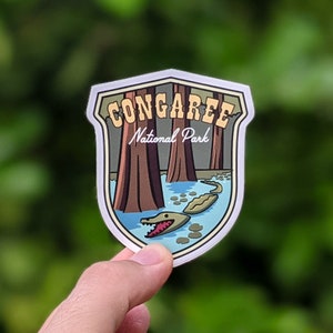 May include: A Congaree National Park sticker with a green alligator in a blue swamp with brown trees and the text "Congaree National Park".
