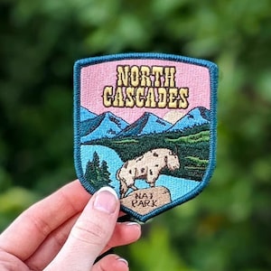 May include: A green and pink embroidered patch with the text "NORTH CASCADES NATIONAL PARK" and a mountain scene with a white mountain goat.