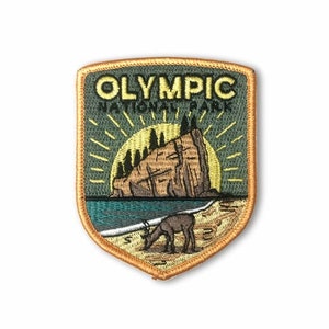 Pode incluir: Um remendo bordado com o logotipo do Parque Nacional Olímpico. O remendo é verde com contorno dourado e representa um pôr do sol sobre um penhasco rochoso com um veado em primeiro plano. O texto "OLYMPIC NATIONAL PARK" é bordado em amarelo.