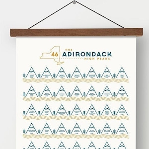 May include: A cream-colored wall art print with a wooden hanger displays a minimalist design. The print features a map of New York State and the text "The Adirondack High Peaks." Below are rows of mountain illustrations and the phrase "The Mountains Are Calling."