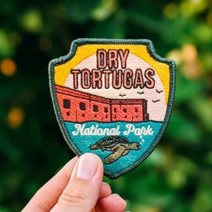 May include: Embroidered patch featuring the text "Dry Tortugas National Park." The patch has a shield shape with a colorful design of a building, a turtle, and a sunset. The patch is held against a blurred green background.