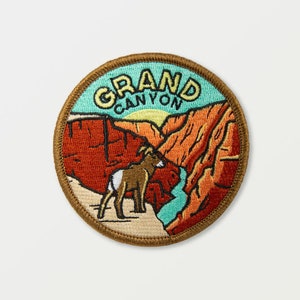 Puede incluir: Parche bordado redondo con un paisaje del Gran Cañón. El parche tiene un borde marrón y las palabras "GRAND CANYON" en amarillo y verde. La imagen muestra formaciones rocosas rojas, un río azul y un sol amarillo, con un borrego cimarrón.