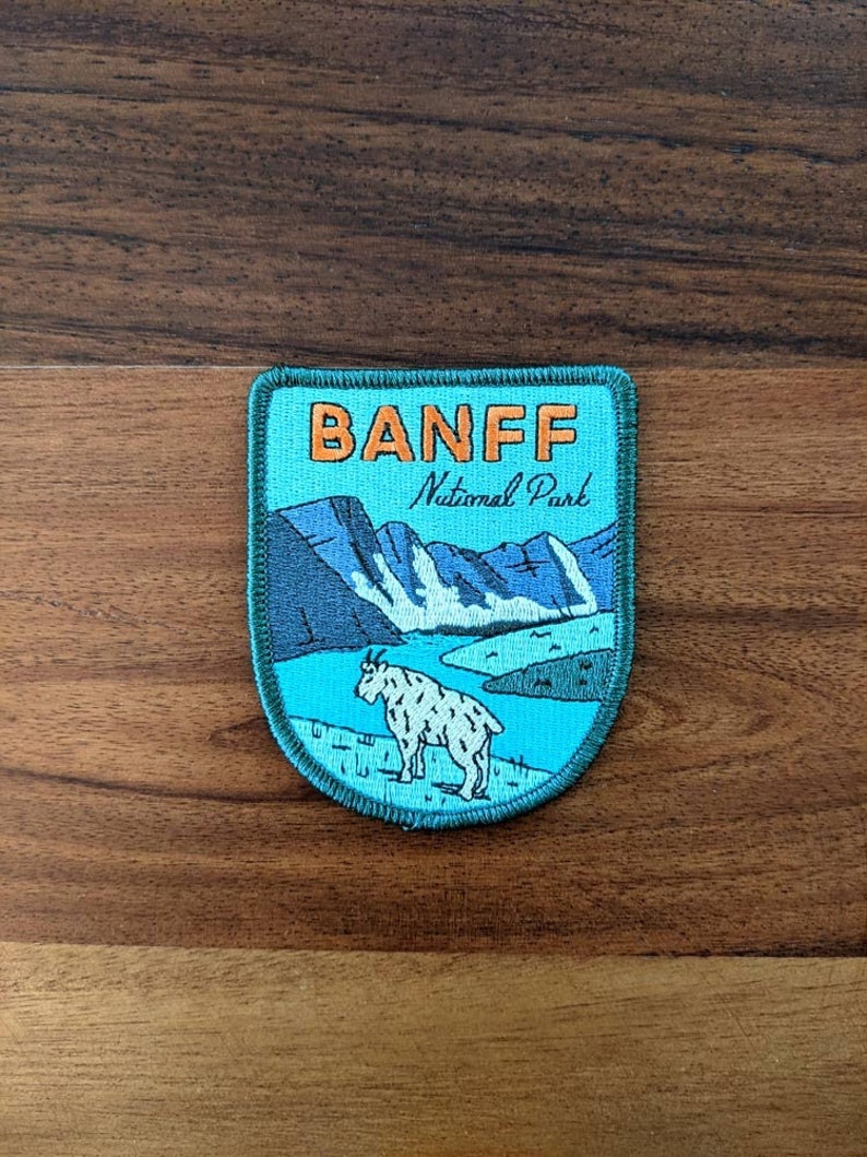 May include: A rectangular patch with a turquoise background features the text "BANFF National Park" in orange and white. The patch depicts a mountain scene with a goat in the foreground. The patch is bordered with a dark blue stitching.