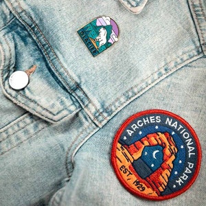 K&ouml;nnte beinhalten: Hellblaue Jeansjacke mit zwei Aufn&auml;hern und einer Anstecknadel. Die Anstecknadel zeigt einen wei&szlig;en B&auml;ren auf gr&uuml;nem Hintergrund mit dem Wort "Glacier". Der runde Aufn&auml;her zeigt "Arches National Park" und "Est. 1929".