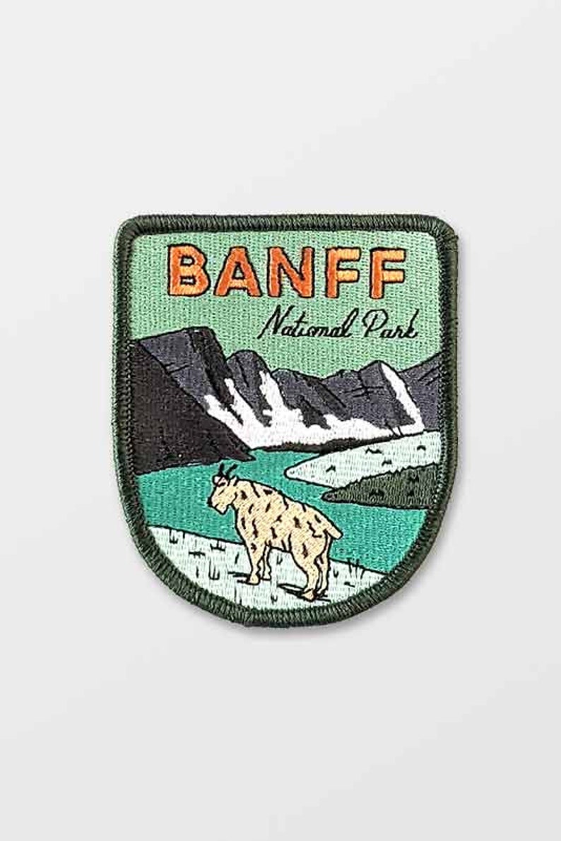 May include: Embroidered patch featuring the text "BANFF National Park" in orange letters. The patch depicts a mountain goat, mountains, and a lake. The patch has a green border and a light green background.