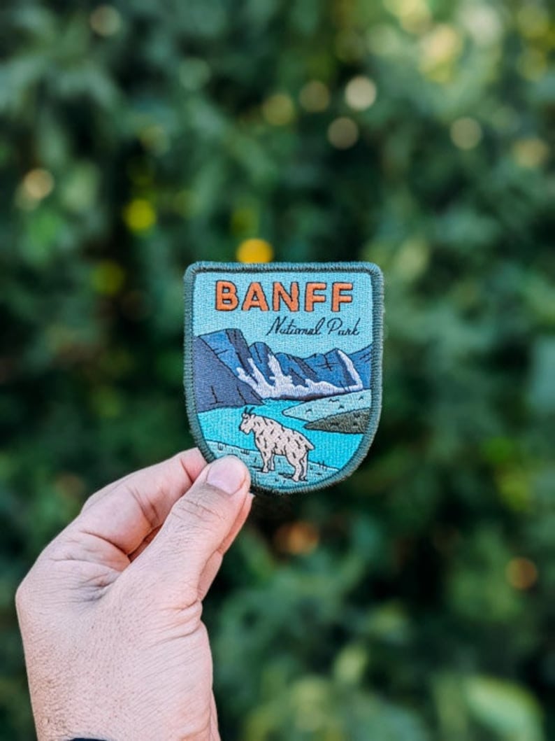 May include: Embroidered patch featuring the text "BANFF National Park" in orange lettering. The patch depicts a mountain goat in front of a lake and mountains. The patch has a blue and green color scheme.