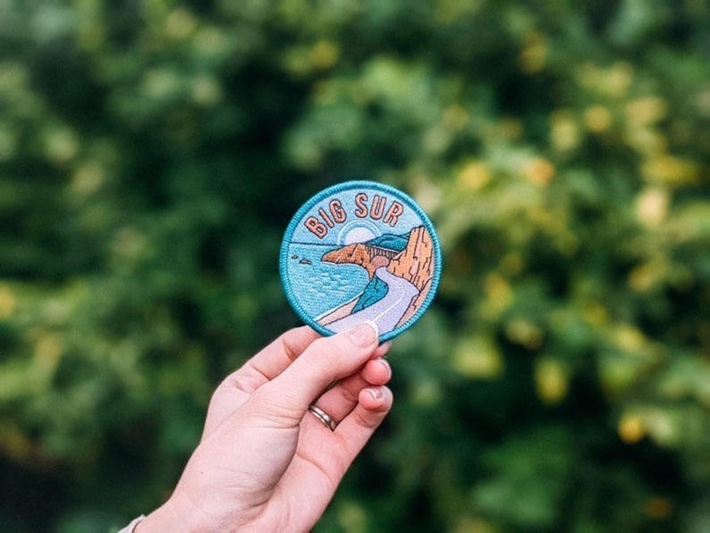 K&ouml;nnte beinhalten: Ein runder Aufn&auml;her mit der Aufschrift "Big Sur" in einem blauen Kreis. Der Aufn&auml;her zeigt eine K&uuml;stenstra&szlig;enszene mit einer Stra&szlig;e, dem Meer und Klippen. Der Aufn&auml;her wird in einer Hand vor einem verschwommenen gr&uuml;nen Hintergrund gehalten.