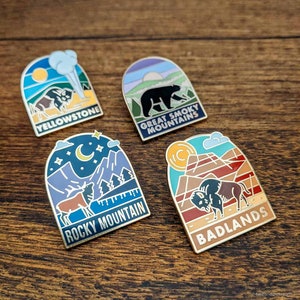 May include: Four enamel pins featuring national park designs. The pins include Yellowstone with a bison, Great Smoky Mountains with a bear, Rocky Mountain with an elk, and Badlands with a bison. Each pin has a gold border.