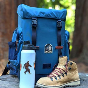 May include: A blue backpack with black straps and buckles, a white water bottle with a Bigfoot sticker, and a tan hiking boot with red laces. The backpack has a Carlsbad patch.