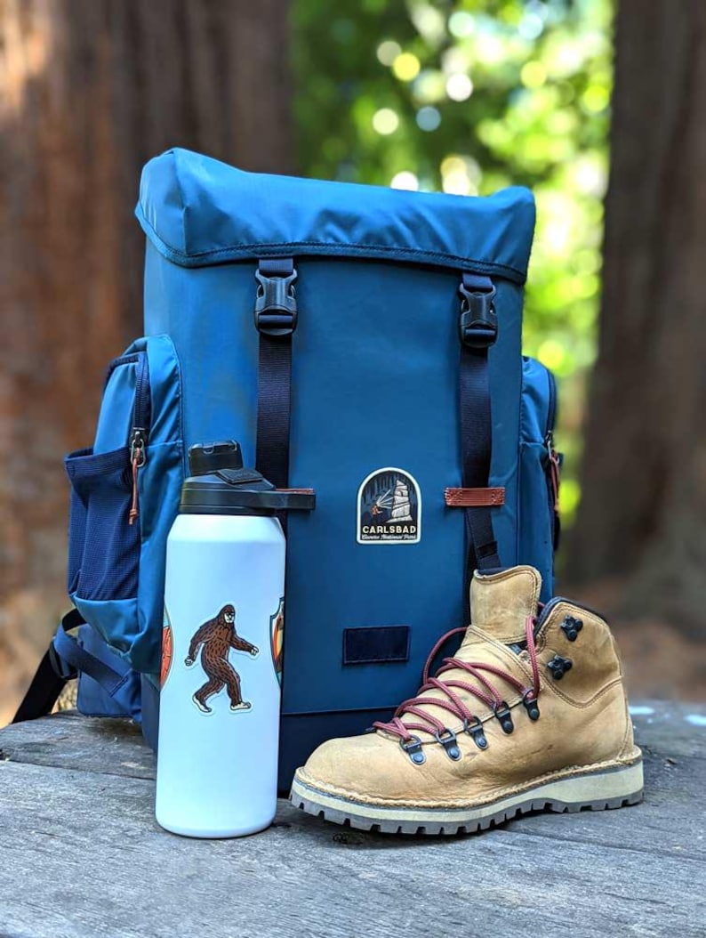 May include: A blue backpack with black straps and buckles, a white water bottle with a Bigfoot sticker, and a tan hiking boot with red laces. The backpack has a Carlsbad patch.