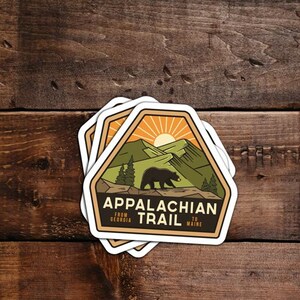 Pu&ograve; includere: Un adesivo con un design marrone, verde e arancione che presenta un orso nero che cammina su un sentiero di montagna. L'adesivo dice "Appalachian Trail" con il testo "From Georgia to Maine" sotto.