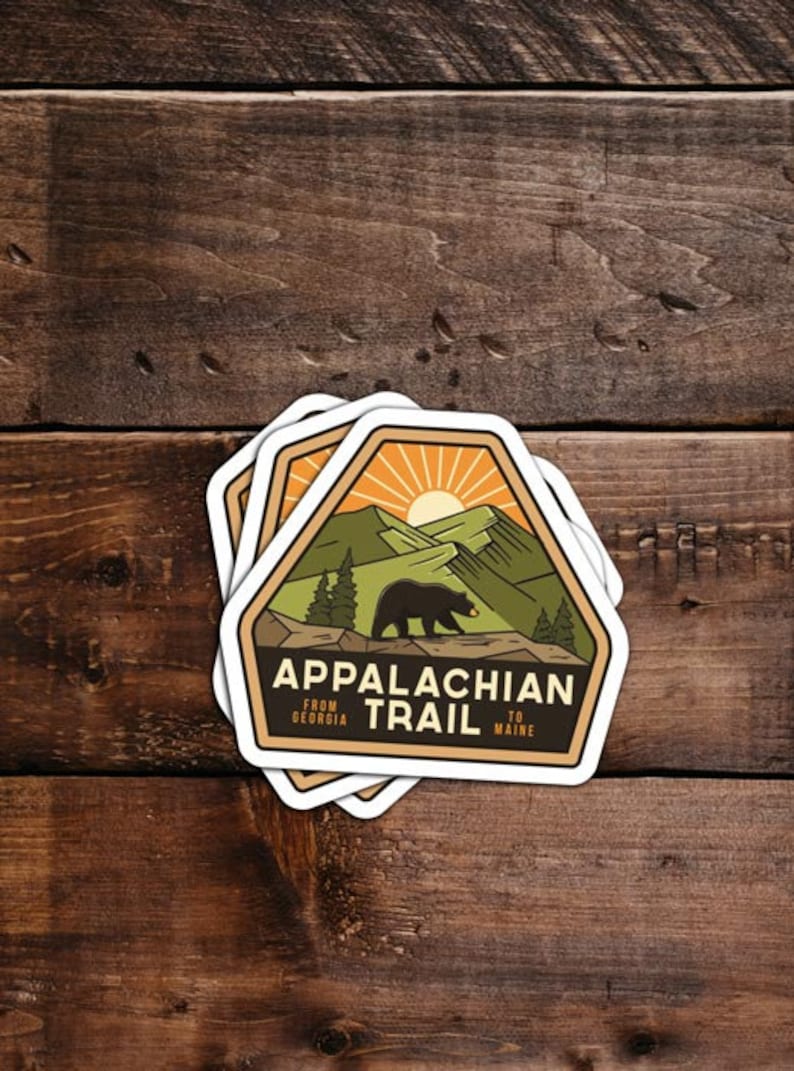 Pu&ograve; includere: Un adesivo con un design marrone, verde e arancione che presenta un orso nero che cammina su un sentiero di montagna. L'adesivo dice "Appalachian Trail" con il testo "From Georgia to Maine" sotto.