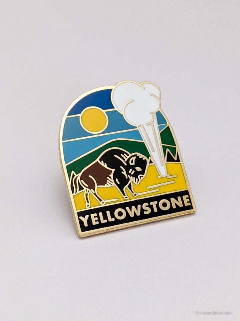 May include: A gold-colored enamel pin featuring a bison in front of a geyser and the text "YELLOWSTONE". The pin has a blue sky with a yellow sun, green hills, and a yellow field.