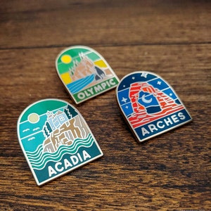 May include: Three enamel pins with arched tops, featuring scenic landscapes. One pin says "OLYMPIC" with a mountain and water scene. Another says "ARCHES" with a red rock formation. The third says "ACADIA" with a coastal scene.