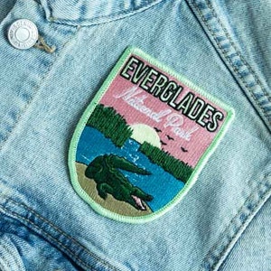 May include: A light blue denim jacket with a green, pink, and blue embroidered patch that says "Everglades National Park". The patch features a green alligator in a river with a pink sunset in the background.