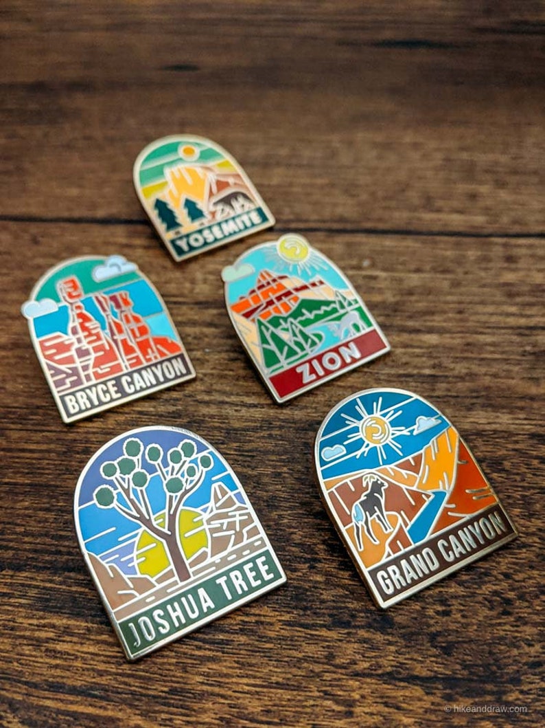 May include: Five enamel pins featuring national park designs. The pins include Yosemite, Bryce Canyon, Zion, Joshua Tree, and Grand Canyon. Each pin has a unique landscape illustration with the park name in a bold font. The pins are set against a wooden background.