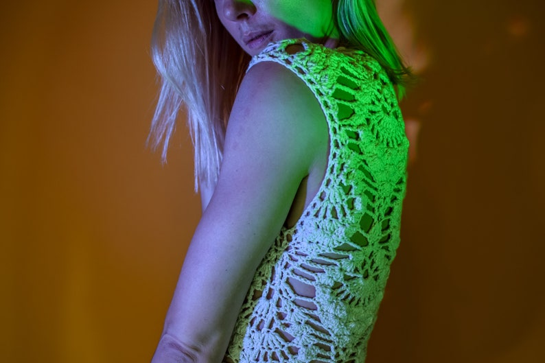 May include: A cream-colored, sleeveless crochet top with a floral pattern. The top has a unique open design and is worn by a person with blonde hair. The background is a gradient of orange and green.