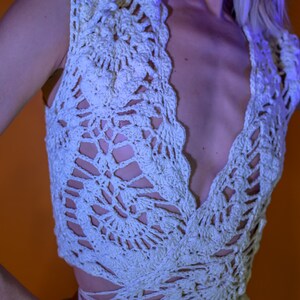 May include: A light cream-colored crochet top with a deep V-neckline. The top features an open, lacy design with a scalloped edge. The garment is sleeveless and appears to be handmade, showcasing intricate patterns.
