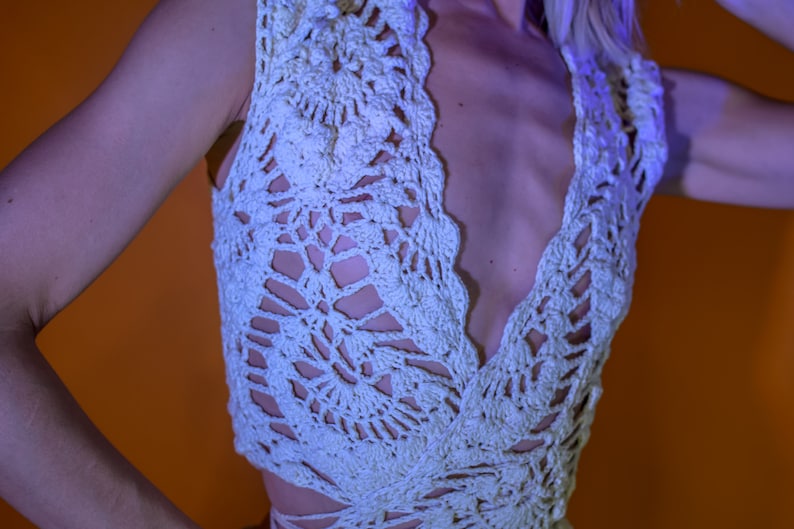 May include: A light cream-colored crochet top with a deep V-neckline. The top features an open, lacy design with a scalloped edge. The garment is sleeveless and appears to be handmade, showcasing intricate patterns.