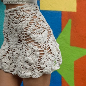 Puede incluir: Falda de ganchillo color crema con dobladillo festoneado. La falda presenta un diseño abierto y de encaje con patrones intrincados. La prenda se muestra sobre un fondo de graffiti colorido, mostrando la textura y el detalle de la pieza hecha a mano.