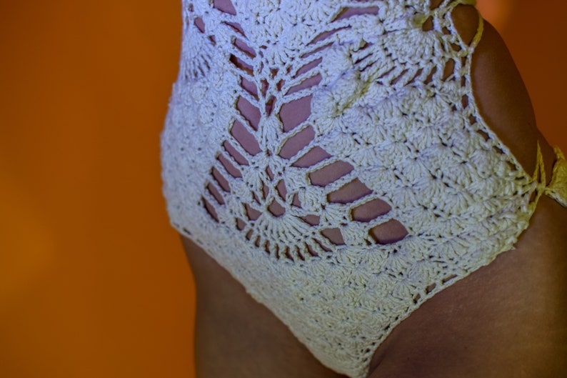 May include: Cream-colored crochet swimsuit with a detailed openwork design. The swimsuit features a high-waisted cut and intricate patterns, showcasing a blend of texture and style. The background is a vibrant orange.
