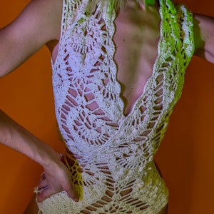 May include: Cream-colored crochet top and matching bottoms. The sleeveless top has a deep V-neck and intricate openwork patterns. The bottoms are high-waisted, also featuring a crochet design. The outfit is set against an orange background.