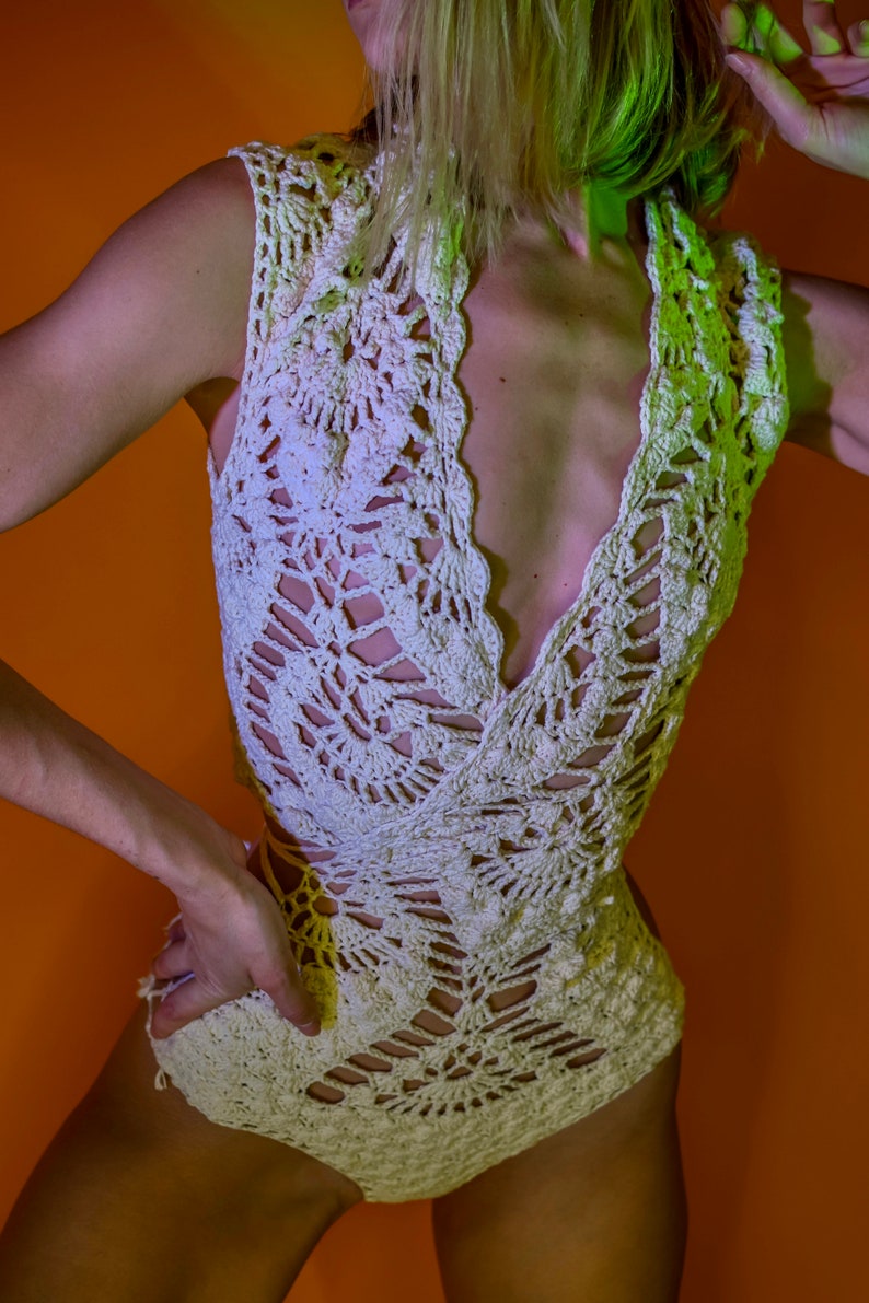 May include: Cream-colored crochet top and matching bottoms. The sleeveless top has a deep V-neck and intricate openwork patterns. The bottoms are high-waisted, also featuring a crochet design. The outfit is set against an orange background.