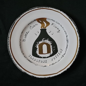 Puede incluir: Plato de cerámica blanco con borde negro y dorado. El plato presenta una ilustración negra de un horno de botella con llamas doradas y el texto "Bottle Oven Firing 78 Gladstone Pottery."