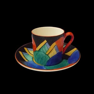 Puede incluir: Un colorido juego de taza y platillo de cerámica. La taza y el platillo son negros con diseños geométricos en rojo, amarillo, azul y verde. La taza tiene un asa roja y un interior color crema.