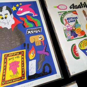 May include: Two colorful illustrations printed on white paper. The first illustration features a vampire with a red cape, a dolphin, a dog, a match, and a safety matchbox with the text "Appu 33" and "Super Deluxe 3". The second illustration features a watermelon, a bowl of noodles, a banjo, a can of Mitsua Cider, and a dragon fruit. The text "Asahi" is printed on the top of the second illustration.