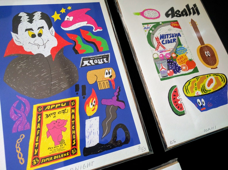 May include: Two colorful illustrations printed on white paper. The first illustration features a vampire with a red cape, a dolphin, a dog, a match, and a safety matchbox with the text "Appu 33" and "Super Deluxe 3". The second illustration features a watermelon, a bowl of noodles, a banjo, a can of Mitsua Cider, and a dragon fruit. The text "Asahi" is printed on the top of the second illustration.