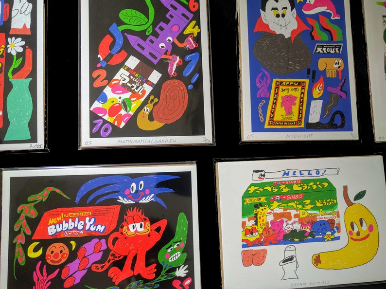 May include: A set of nine colorful prints featuring various illustrations, including a mathematical garden, a vampire, a cartoon cat, and a pear. The prints are framed in black and have white borders with text at the bottom.